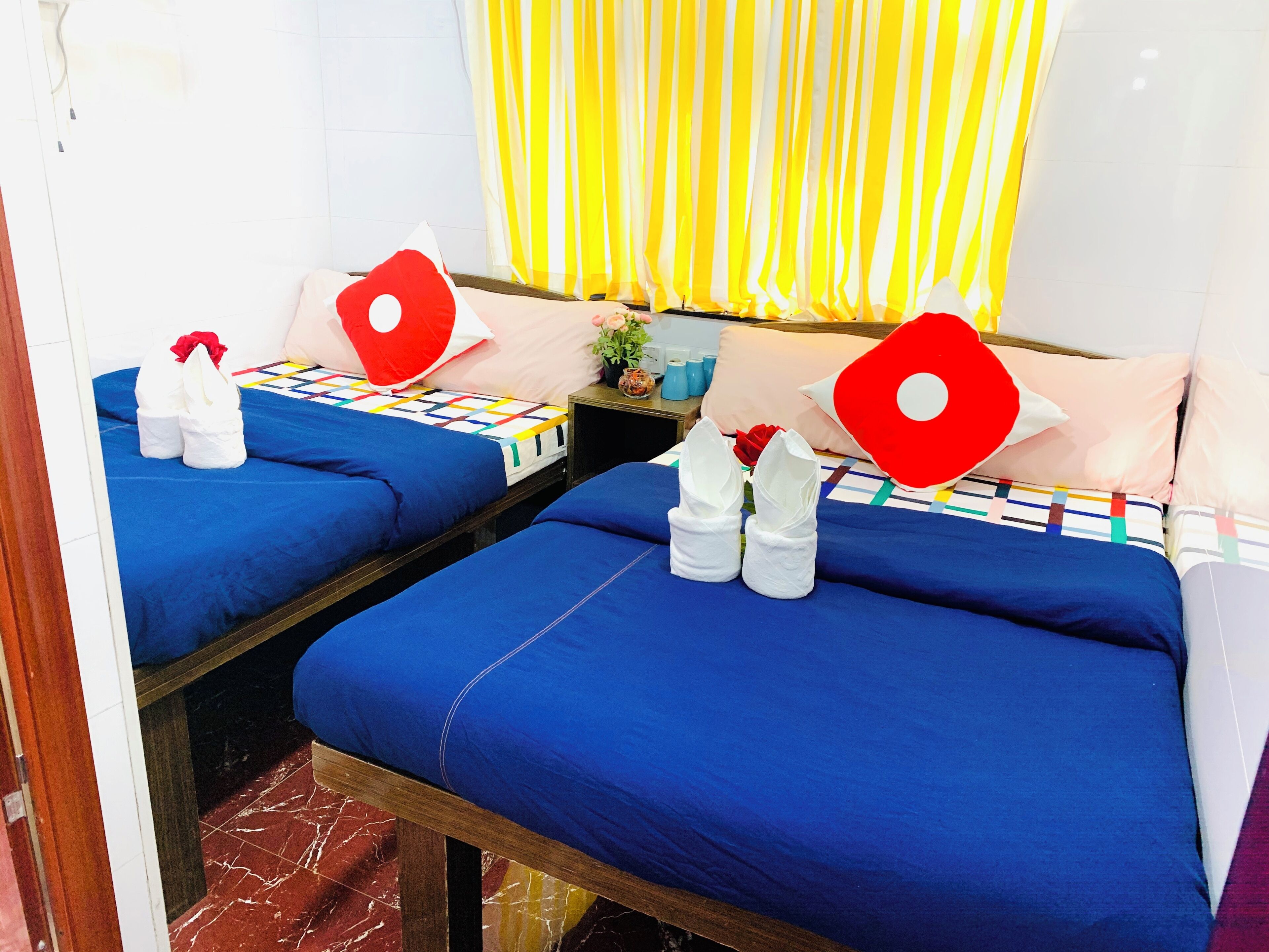 A hotel room with two beds, each with blue bedding and white towels. There are red heart-shaped pillows and a small table with a vase of flowers.