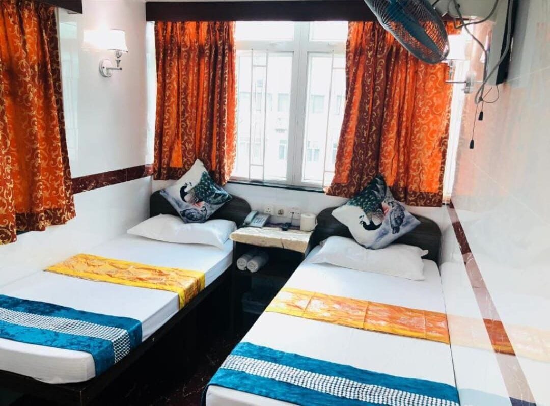 Ein Zimmer mit zwei Betten, jeweils mit gemusterten Bettdecken und Kissen, einem kleinen Tisch mit Telefon und einem Fenster mit Vorhängen.