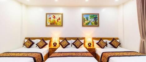 Ein Hotelzimmer mit drei hölzernen Betten, jeweils mit weißer Bettwäsche und Zierkissen. An den Wänden hängen zwei Gemälde, eines zeigt Blumen und das andere eine Landschaft. Die Zimmerdecke ist mit violetter Beleuchtung ausgestattet.