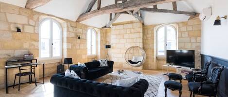 A spacious living room with exposed wooden beams, stone walls, and large windows.