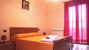 Un dormitorio con cama, mesita de noche, lámpara, radiador y ventana con cortinas rojas.