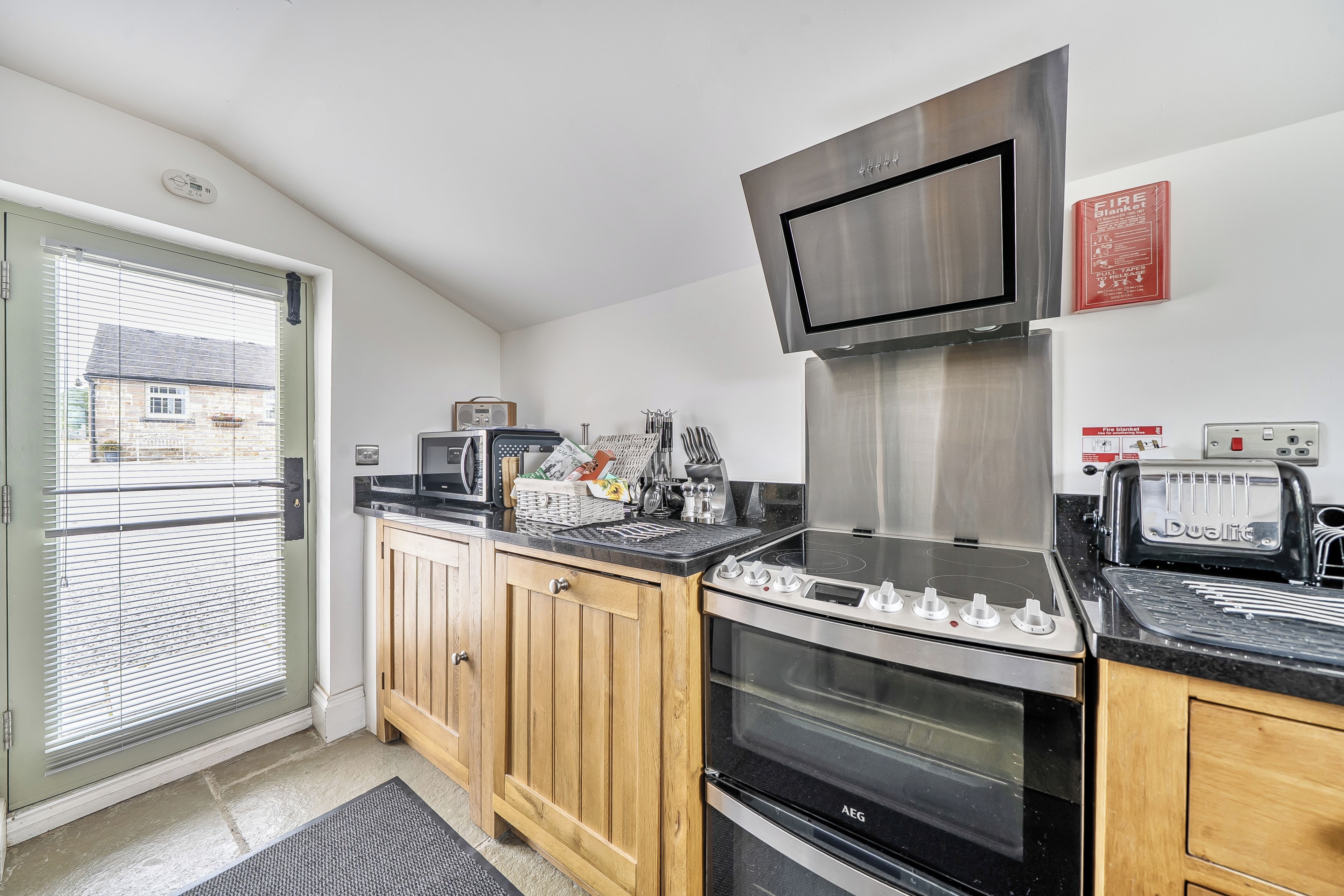 A compact kitchen with a built-in oven, a stove, and a microwave. There is a window with blinds, a door, and a fire safety sign on the wall.