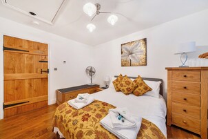 A hotel room with a bed, bedside table, lamp, fan, artwork, and wooden wardrobe.