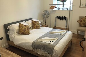 Ein ordentlich bezogenes Bett mit gemusterten Kissen, ein Ventilator am Nachttisch und ein kleiner Tisch mit einer Vase und Dekoration.