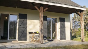 A covered patio area with a pergola structure, large sliding glass doors, and a small table with a chair. - Maison La Fontaine - Paisible & Verdoyante (Trélazé)