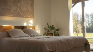 A bedroom with a large bed, a nightstand with a lamp, a world map on the wall, and a view of the outdoors through a window. - Maison La Fontaine - Paisible & Verdoyante (Trélazé)