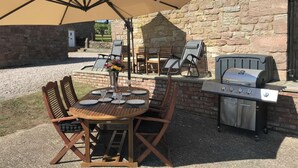 Un patio al aire libre con una mesa redonda de madera, cuatro sillas, una sombrilla color crema, una parrilla y un muro de piedra al fondo.