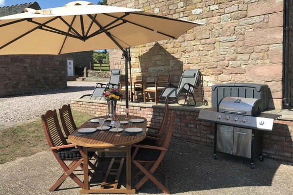 An outdoor patio with a round wooden table, four chairs, a beige umbrella, a grill, and a stone wall in the background.