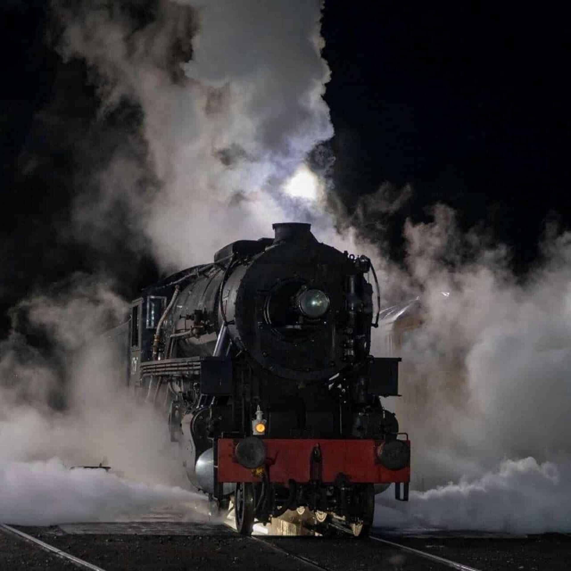 A steam locomotive with a prominent front headlight and a red buffer beam, emitting steam, on a track.