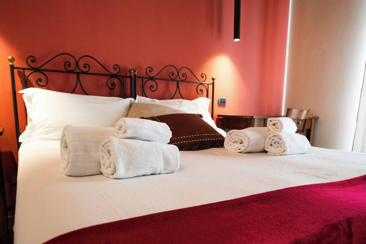 Una cama bien hecha con sábanas blancas y toallas enrolladas, cabecera de metal oscuro y pared de acento roja.