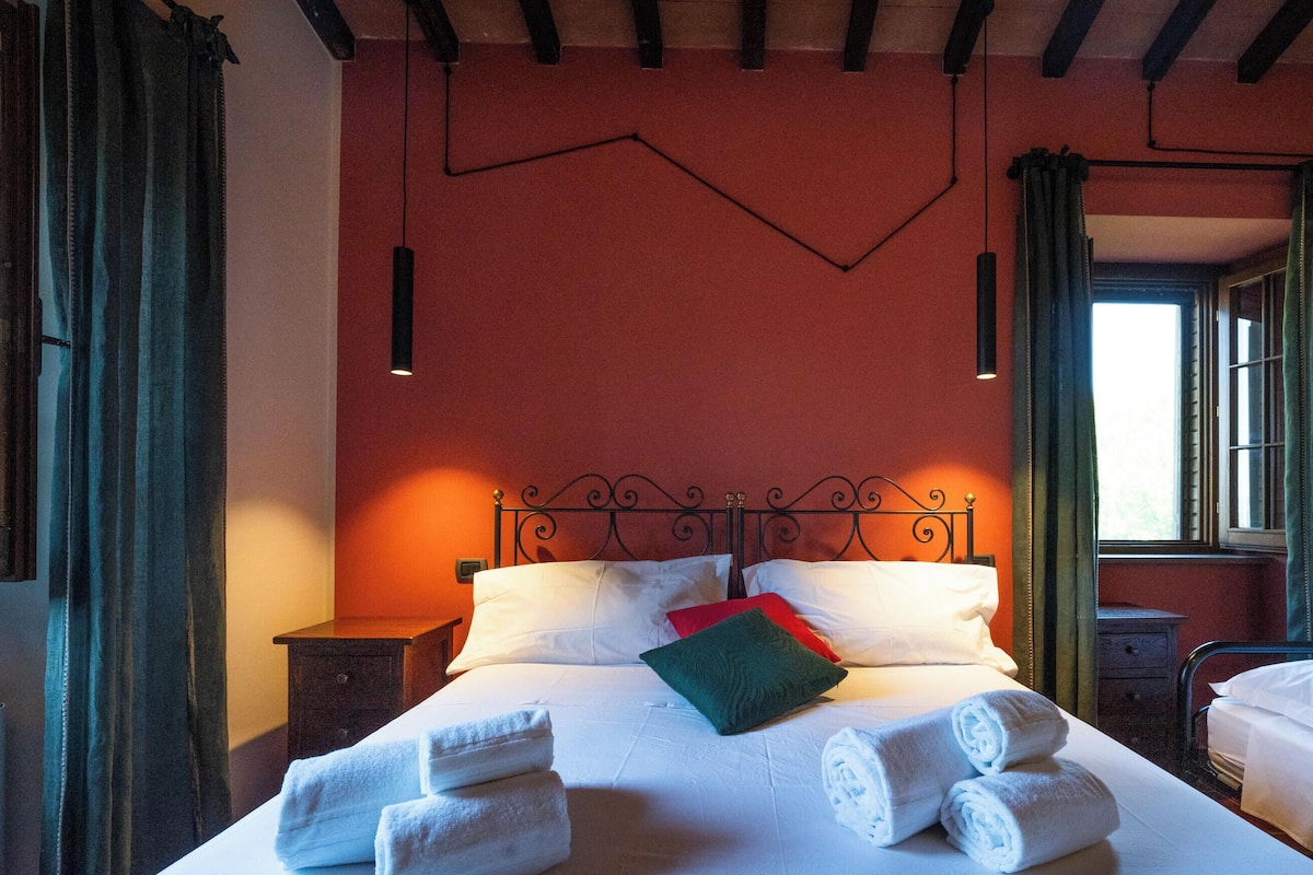 Una cama bien hecha con sábanas blancas y toallas enrolladas, un cojín decorativo rojo y un cabecero de metal. Hay dos mesitas de noche, una con una lámpara, y una ventana con cortinas verdes.
