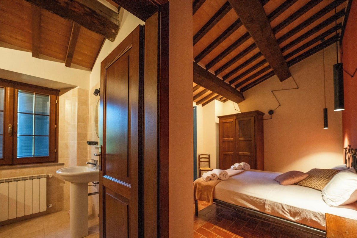 Un dormitorio con cama, mesita de noche y cabecero de madera. Hay una ventana con persianas y un radiador en el baño contiguo.