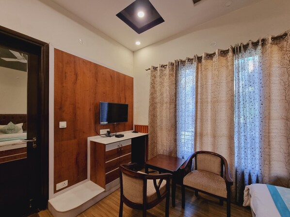 A hotel room with a wooden wardrobe, a desk with a television, two chairs, a bed, and a window with curtains. - Stylia Suites (Dalhousie)