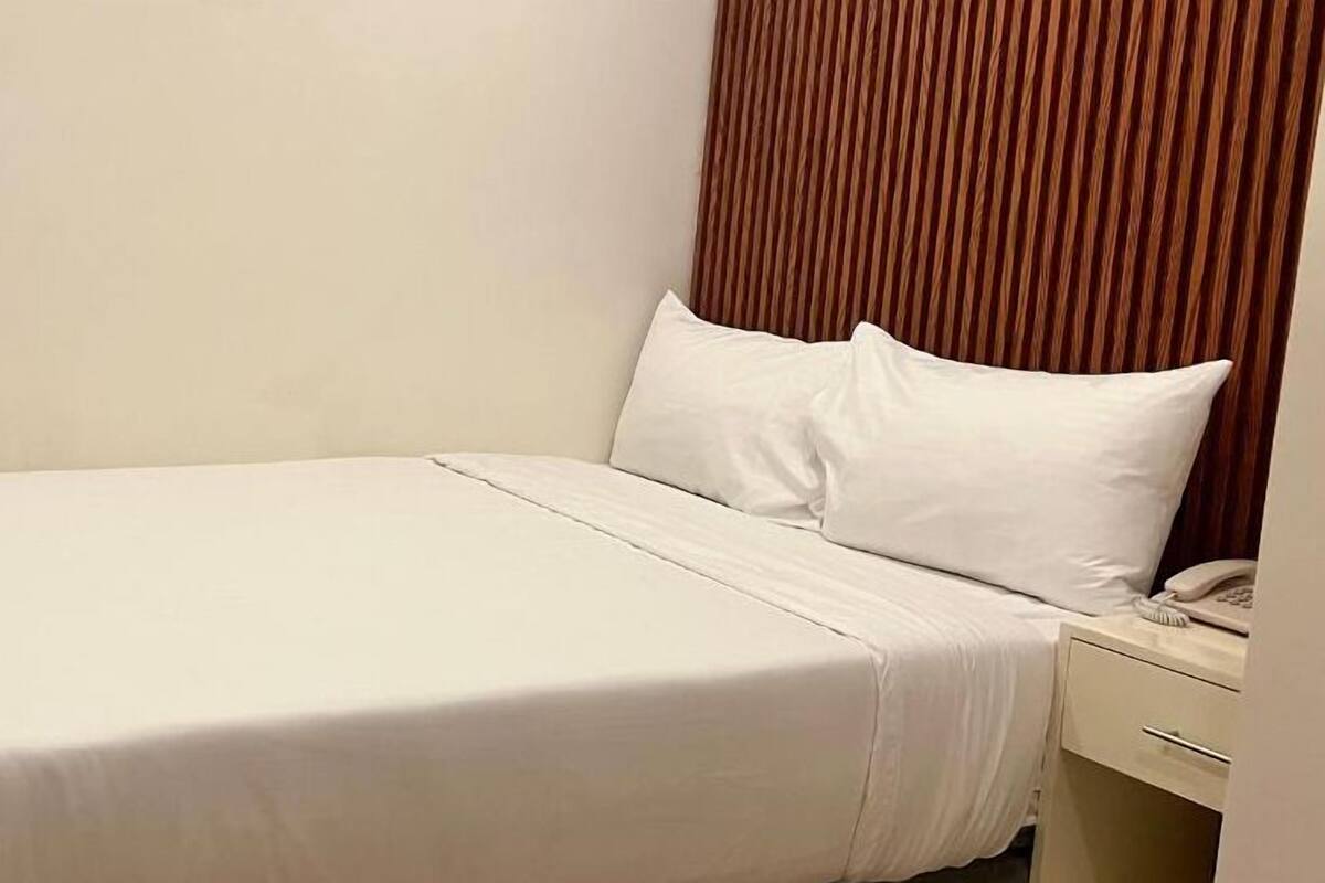 A small, neatly made bed with white bedding in a simple room with a wooden headboard and a small bedside table with a phone.