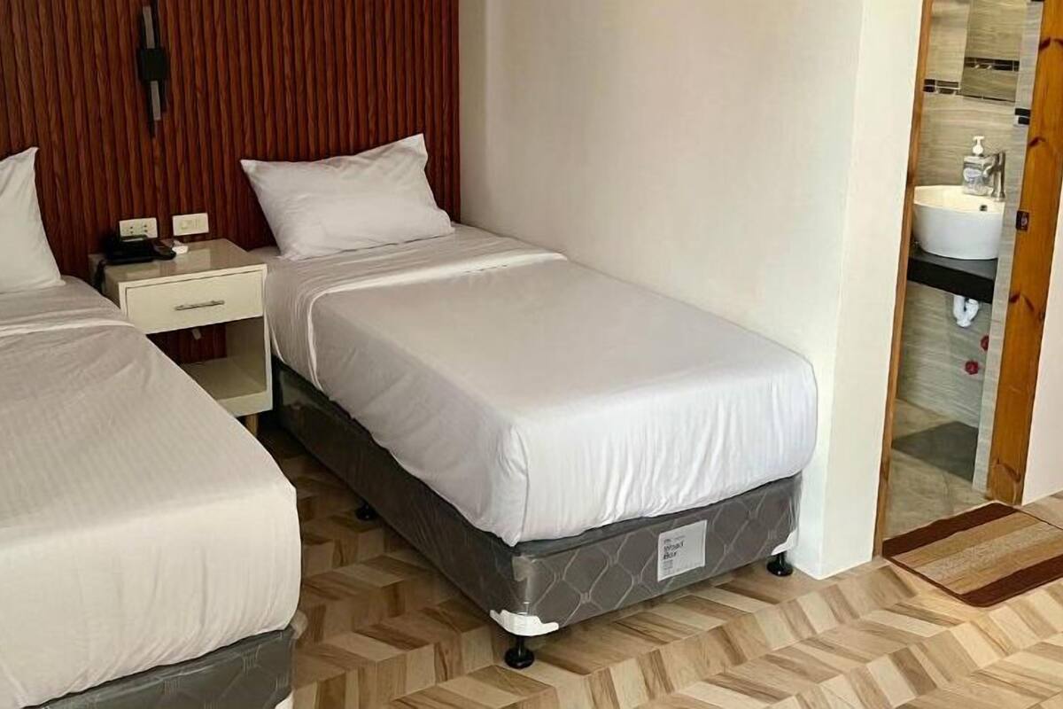 A hotel room with two single beds, a wooden headboard, a nightstand, a small table, and a bathroom visible through an open door.