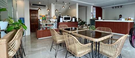 A modern hotel lobby with a central dining area featuring wicker chairs and a glass-top table. There is a reception desk with a counter and a person behind it.