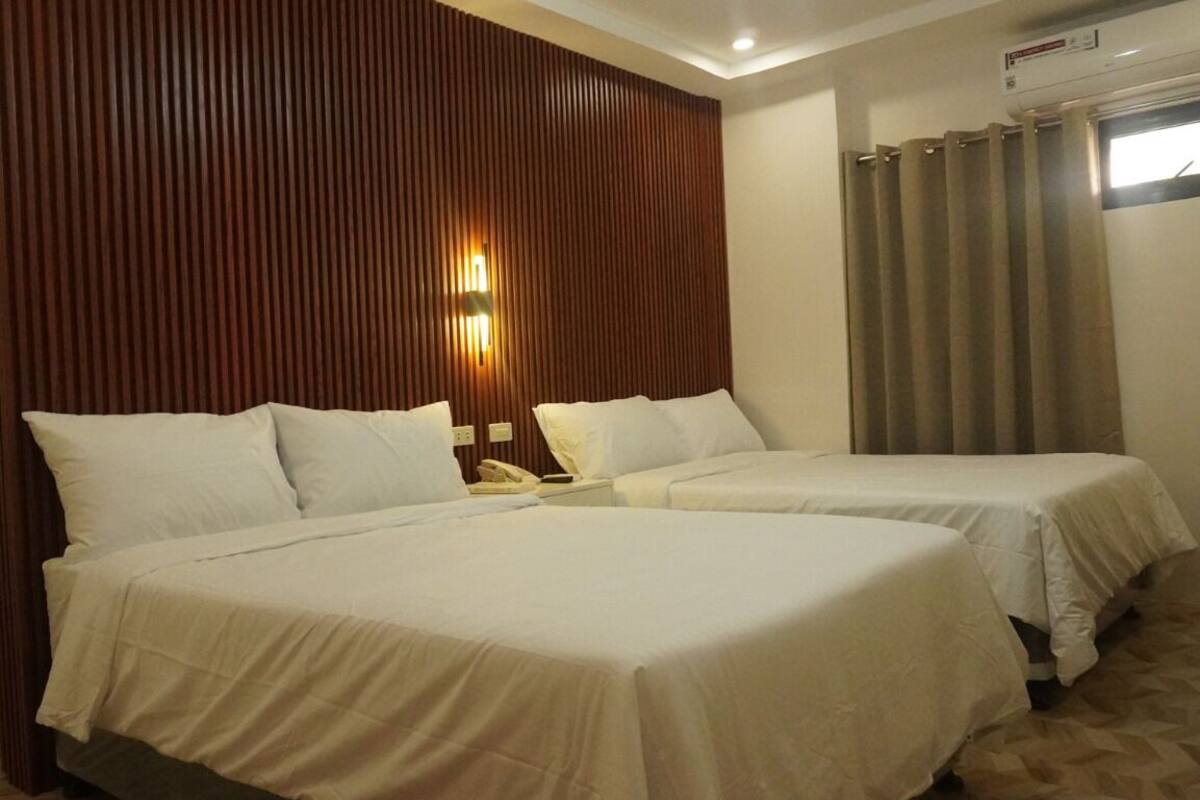 A hotel room with two beds, a wooden panel wall, a wall-mounted phone, and a window with curtains.