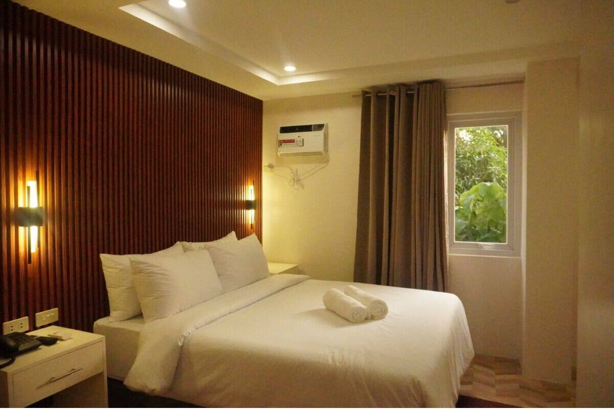 A hotel room with a bed, bedside table, lamp, air conditioning unit, window with curtains, and a view of greenery outside.