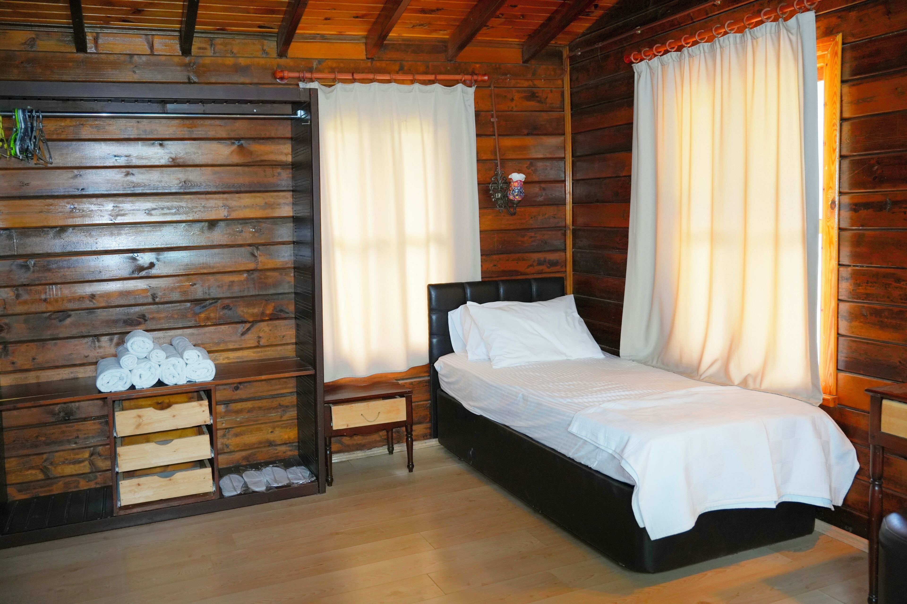 Una habitación tipo cabaña de madera con cama, cortinas y una mesita de noche de madera.