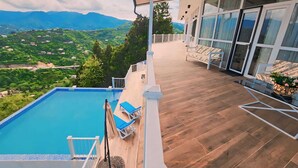 Una piscina sul tetto con un solarium in legno, un'area relax con divano e poltrone, e vista su montagne e vegetazione.