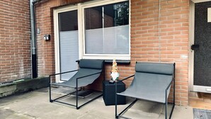 Un patio con dos sillas de metal, una pequeña mesa, un muro de ladrillo y una ventana con persianas.