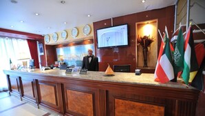 A hotel reception desk with clocks, a TV, flags, and a man in a suit. - Hôtel Abbasside Palace - Palm Beach (Algiers)