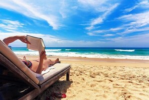 A person lying on a beach chair reading a book, with a wooden bench and sandals nearby. - Hôtel Abbasside Palace - Palm Beach (Algiers)