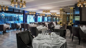 Een restaurant met ronde tafels gedekt voor diners, grote ramen en moderne kroonluchters.