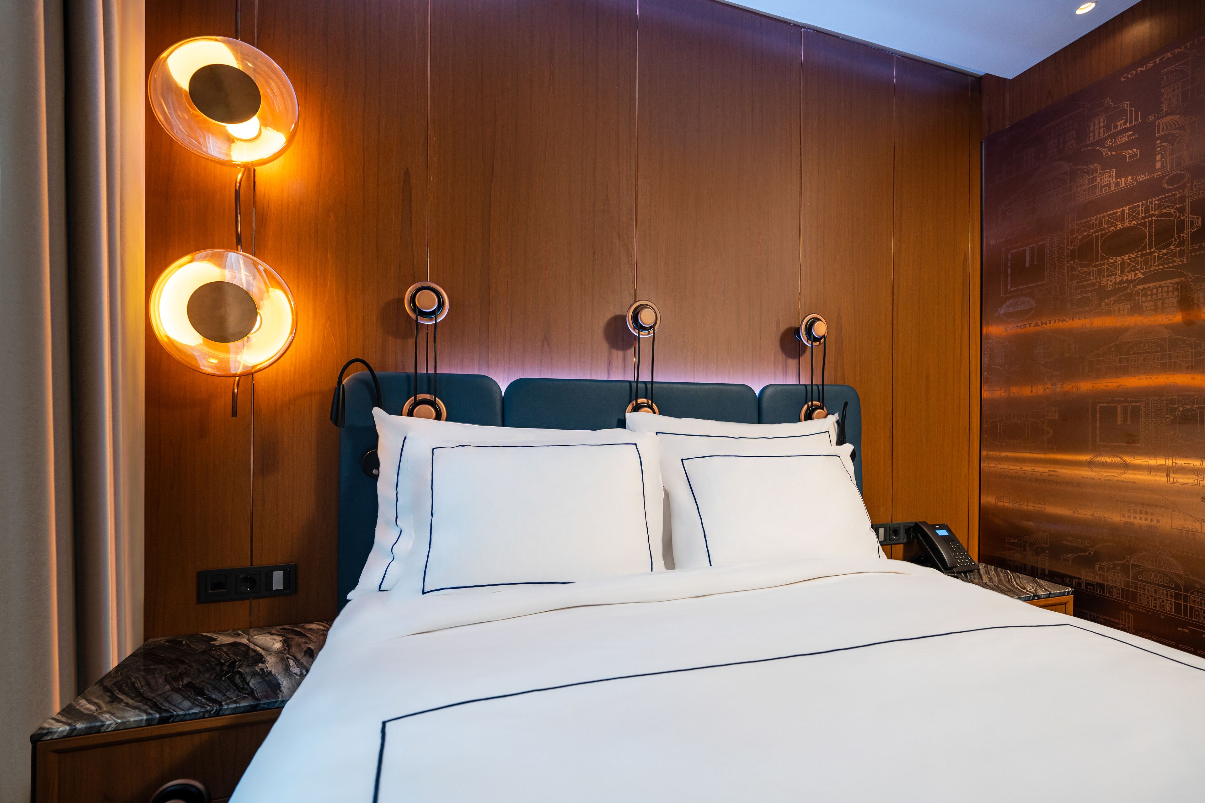 Habitación de hotel con una cama grande, dos mesitas de noche, un cabecero de madera y una pared con un mural de un paisaje urbano.