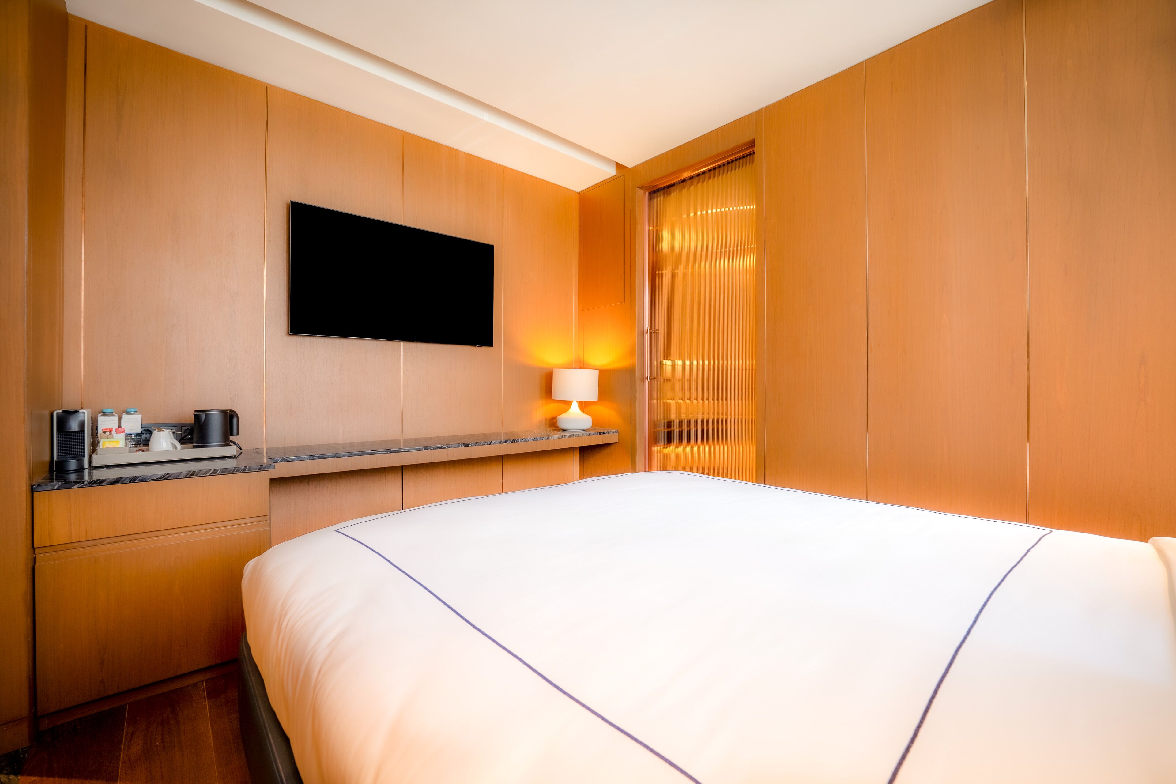 A hotel room with a large bed, a flat-screen TV mounted on the wall, a bedside table with a lamp, and a wooden wardrobe.