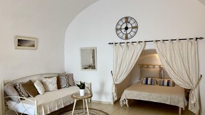 A cozy room with a sofa, a small table with a plant, a mirror, and a bed with curtains. - Santorini Cave Escape in Oia w Patio (Santorini)