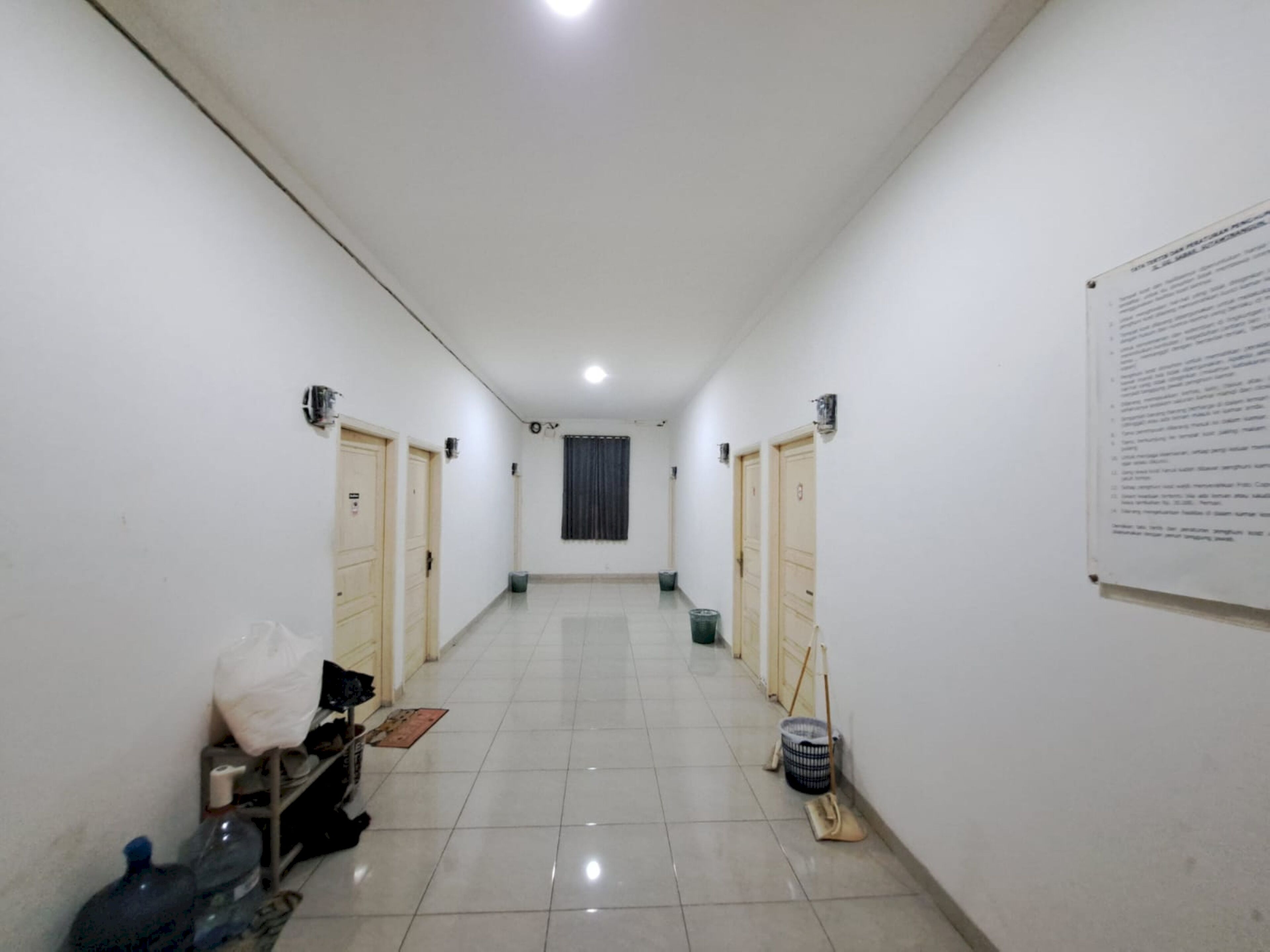 A corridor with white walls, tiled floor, and doors on both sides. There is a notice board on the right wall and cleaning supplies on the floor.