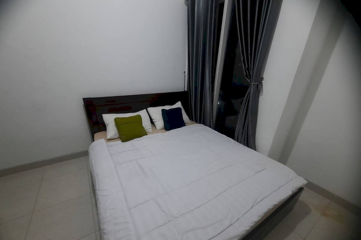 A bed with a white mattress and pillows in various colors, a dark headboard, and a window with grey curtains.