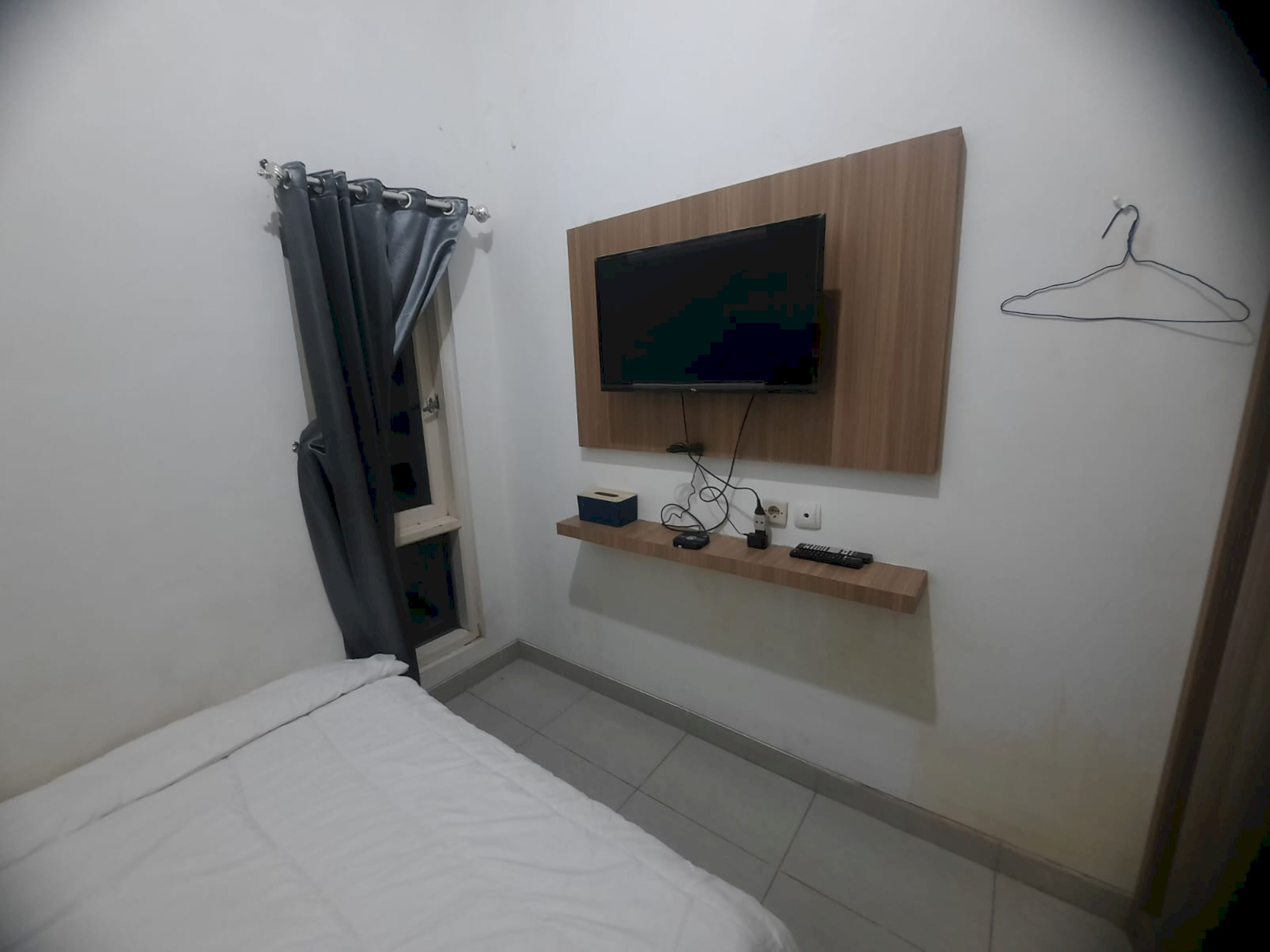 A small, neatly arranged bedroom with a bed, a television mounted on the wall, a small shelf with a remote control, and a hanger on the wall.