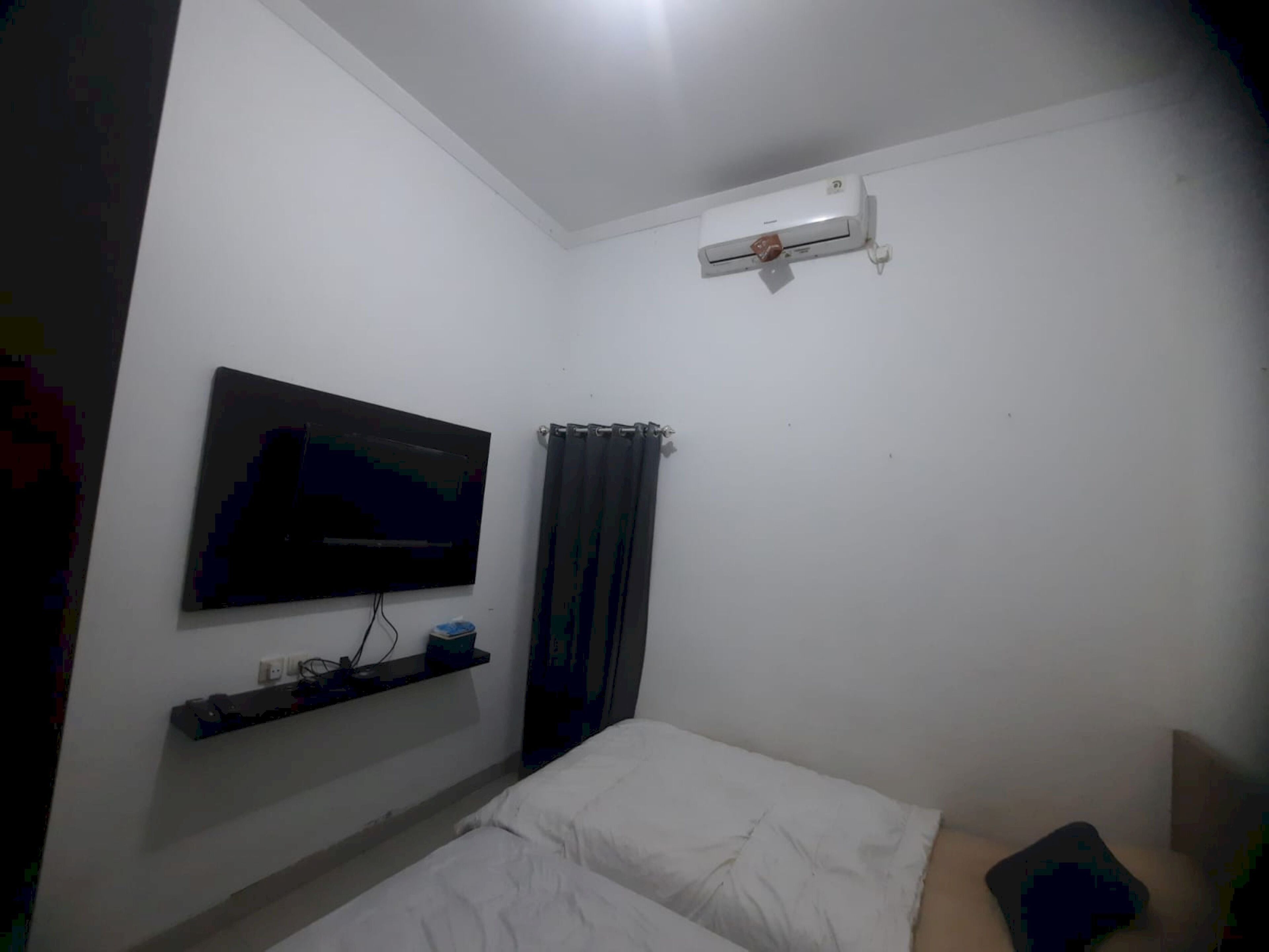 A small, sparsely furnished room with a single bed, a mounted TV, a wall-mounted air conditioner, and a curtain.
