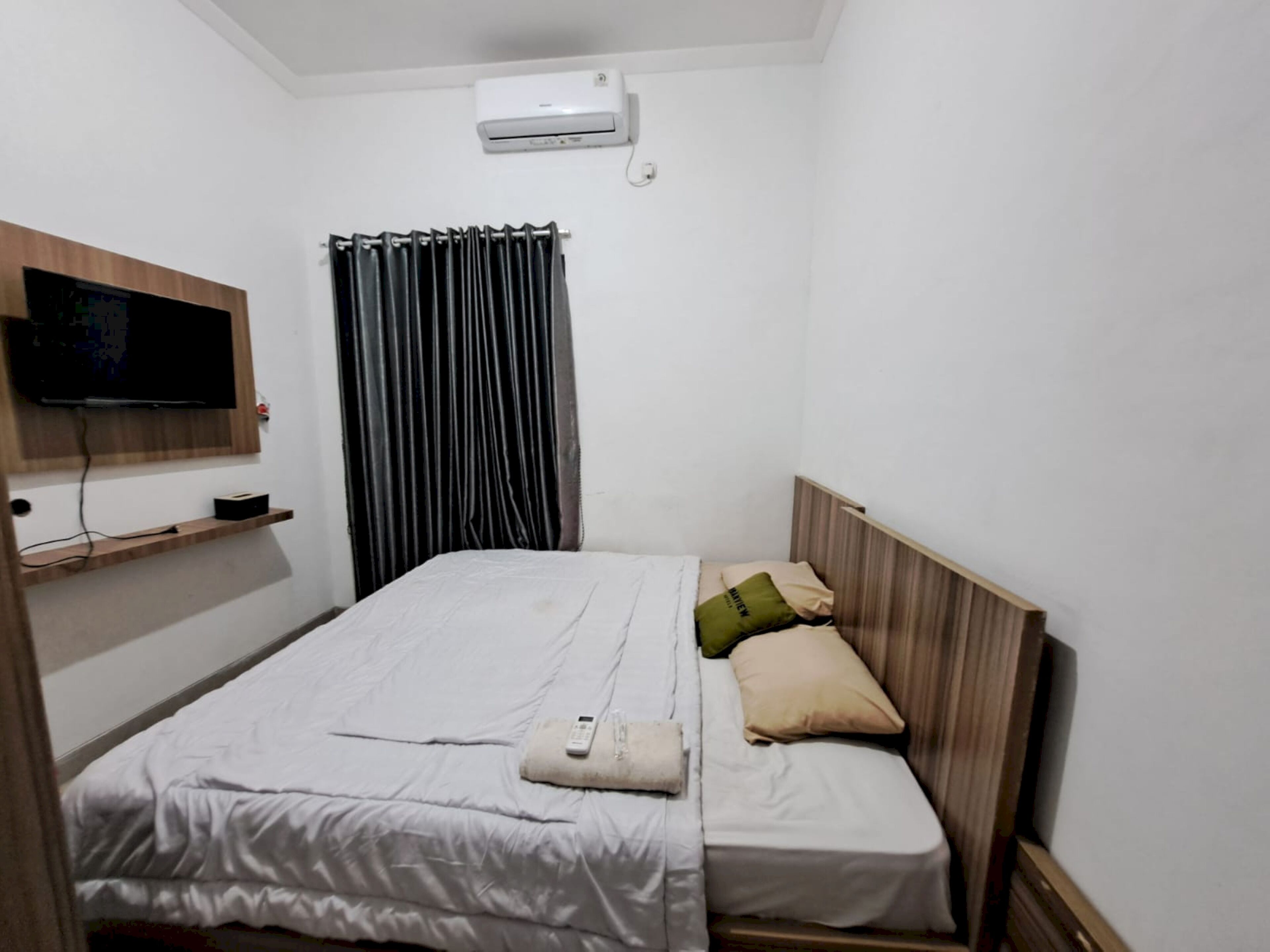 Een kleine, verzorgde kamer met een bed, een wandgemonteerde televisie, een plank en een raam met gordijnen.