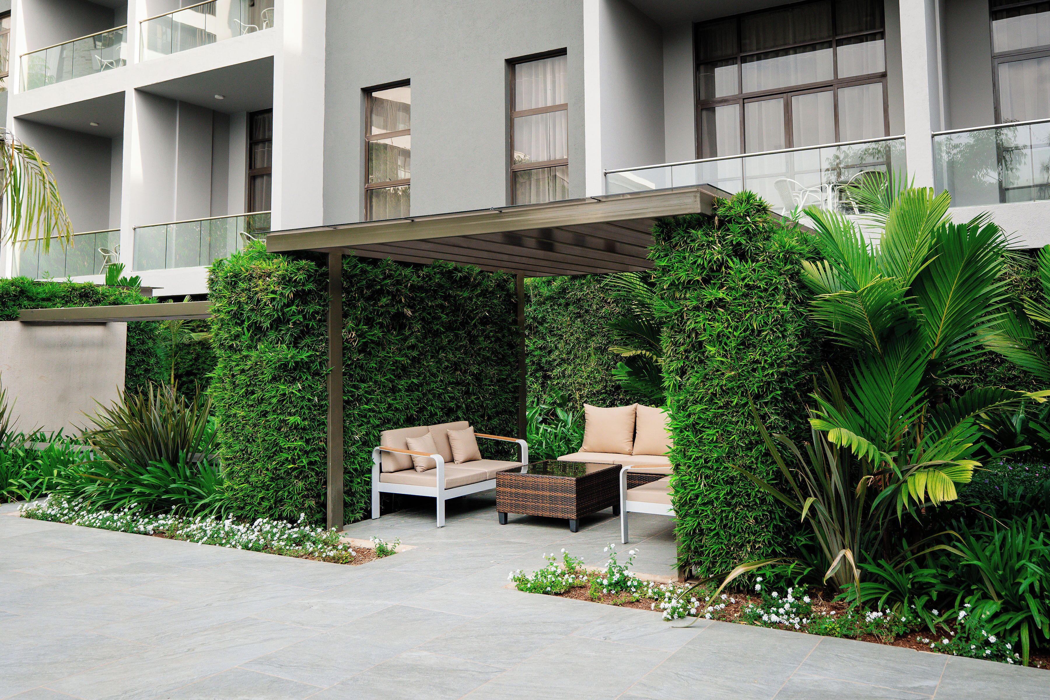 A modern outdoor seating area with a sofa and a coffee table, surrounded by lush greenery and a concrete floor.