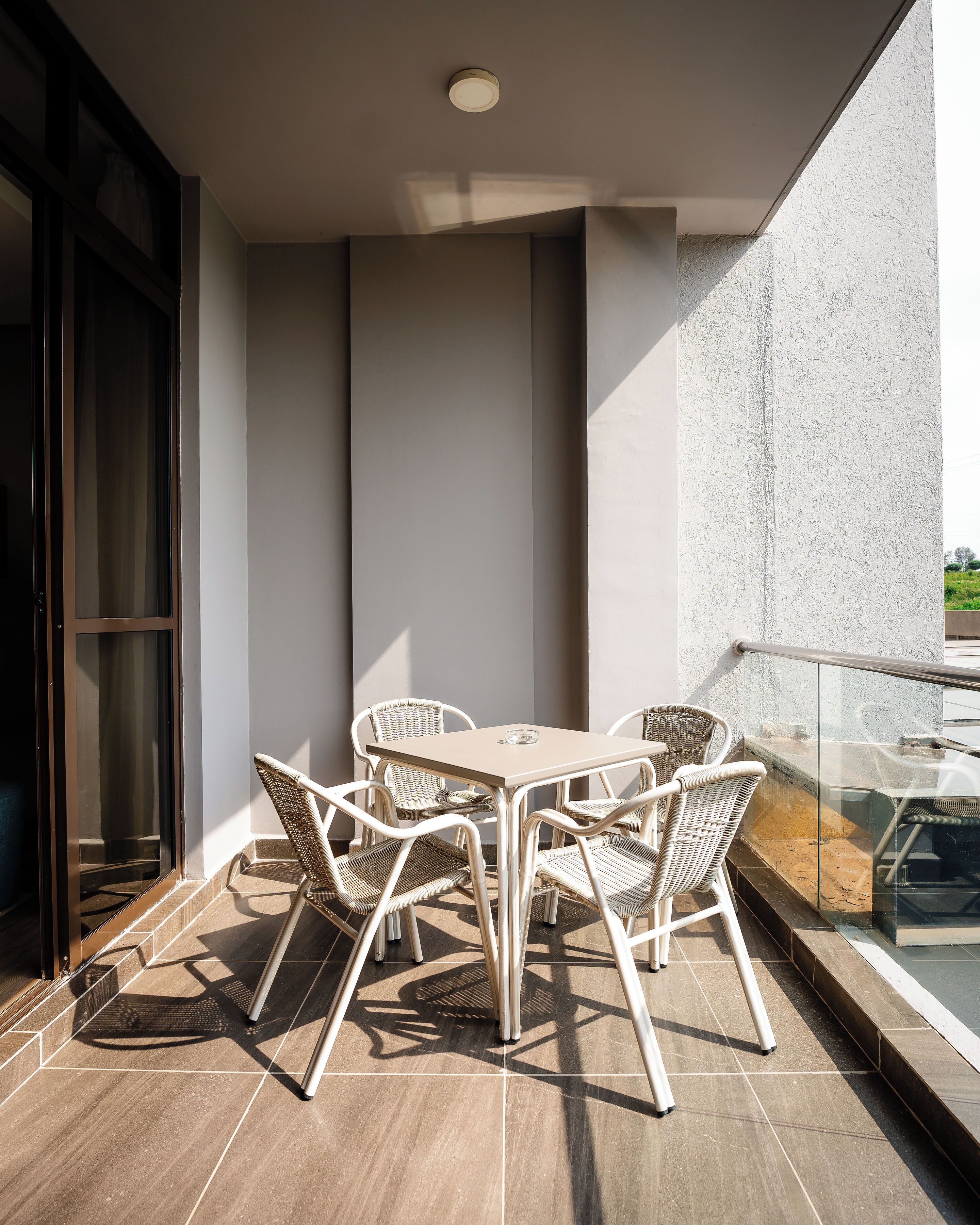 A balcony with a table and four wicker chairs, a glass railing, and a view of greenery.