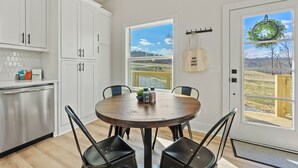 A modern kitchen with white cabinets, a stainless steel dishwasher, and a round wooden table with four chairs. There are windows with views of a landscape and a wreath hanging on one. - War Eagle Creek Retreat- Cabin on the Creek- Private but close to everything (HUNTSVILLE)