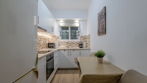 A compact kitchen with white cabinets, a microwave, a toaster, and a coffee maker. There is a window with a view of greenery outside, and a small table with a plant on it. - Kalithea Gardenspot (Kallithea)
