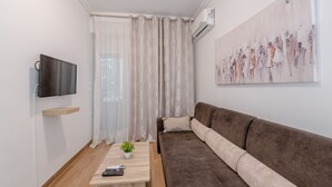 A modern living room with a brown sofa, a small table with a plant, a mounted TV, and a large abstract painting on the wall. - Kalithea Gardenspot (Kallithea)