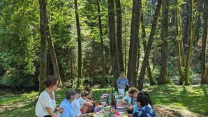 Eine Gruppe von Personen speist an einem Holztisch im Freien in bewaldeter Landschaft.