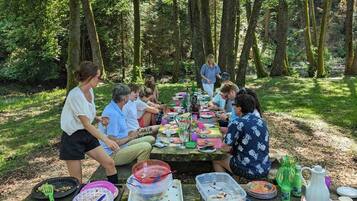 Un groupe de personnes est assis à une table en bois, à l’extérieur, dans un endroit boisé, et y prend son repas.