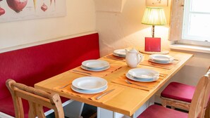 Ein Essbereich mit einem Holztisch, einer rot gepolsterten Bank, Holzsesseln, einer Lampe und einem an der Wand hängenden Bild mit Gemüse.