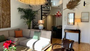 Ein modernes Wohnzimmer mit Sofa, Couchtisch und einem Sessel. Eine Treppe mit Geländer, eine Pflanze und eine Lampe sind ebenfalls zu sehen.