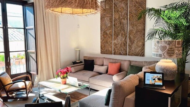 Ein modernes Wohnzimmer mit Sofa, Couchtisch und einem Laptop auf einem Beistelltisch. Ein großes Fenster mit Vorhängen, eine Pflanze und eine dekorative Wandverkleidung sind ebenfalls vorhanden.