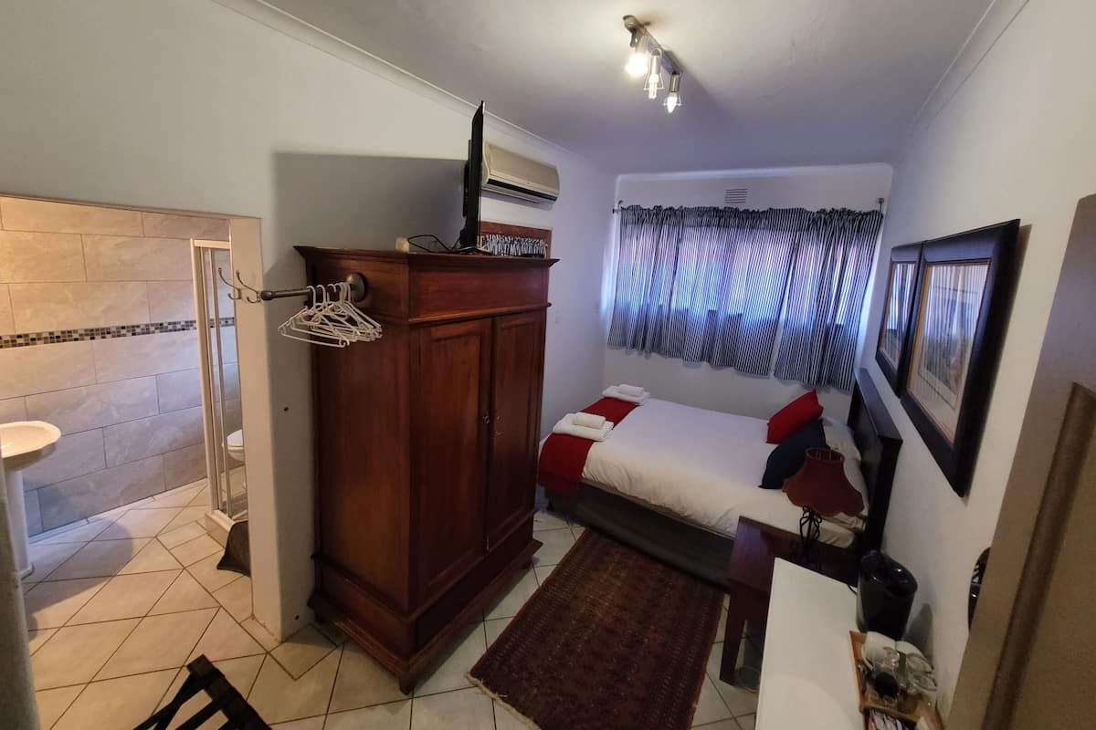 Habitación de hotel compacta con ducha, armario de madera, cama con almohada roja, alfombra pequeña e iluminación en el techo.