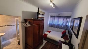 A compact hotel room with a shower, a wooden wardrobe, a bed with a red pillow, a small rug, and a ceiling light fixture.