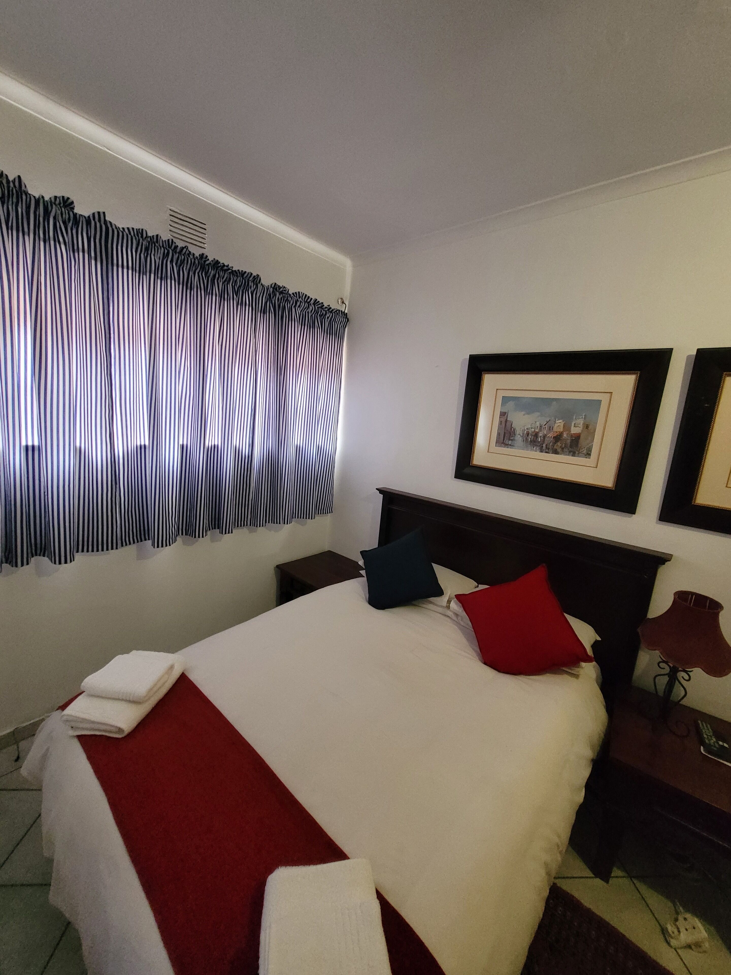 Quarto de hotel com uma cama bem arrumada, uma pequena mesa de cabeceira com uma luminária e uma janela com cortinas listradas.