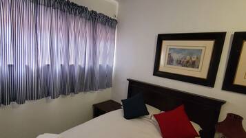 Quarto de hotel com uma cama bem arrumada, uma pequena mesa de cabeceira com uma luminária e uma janela com cortinas listradas.